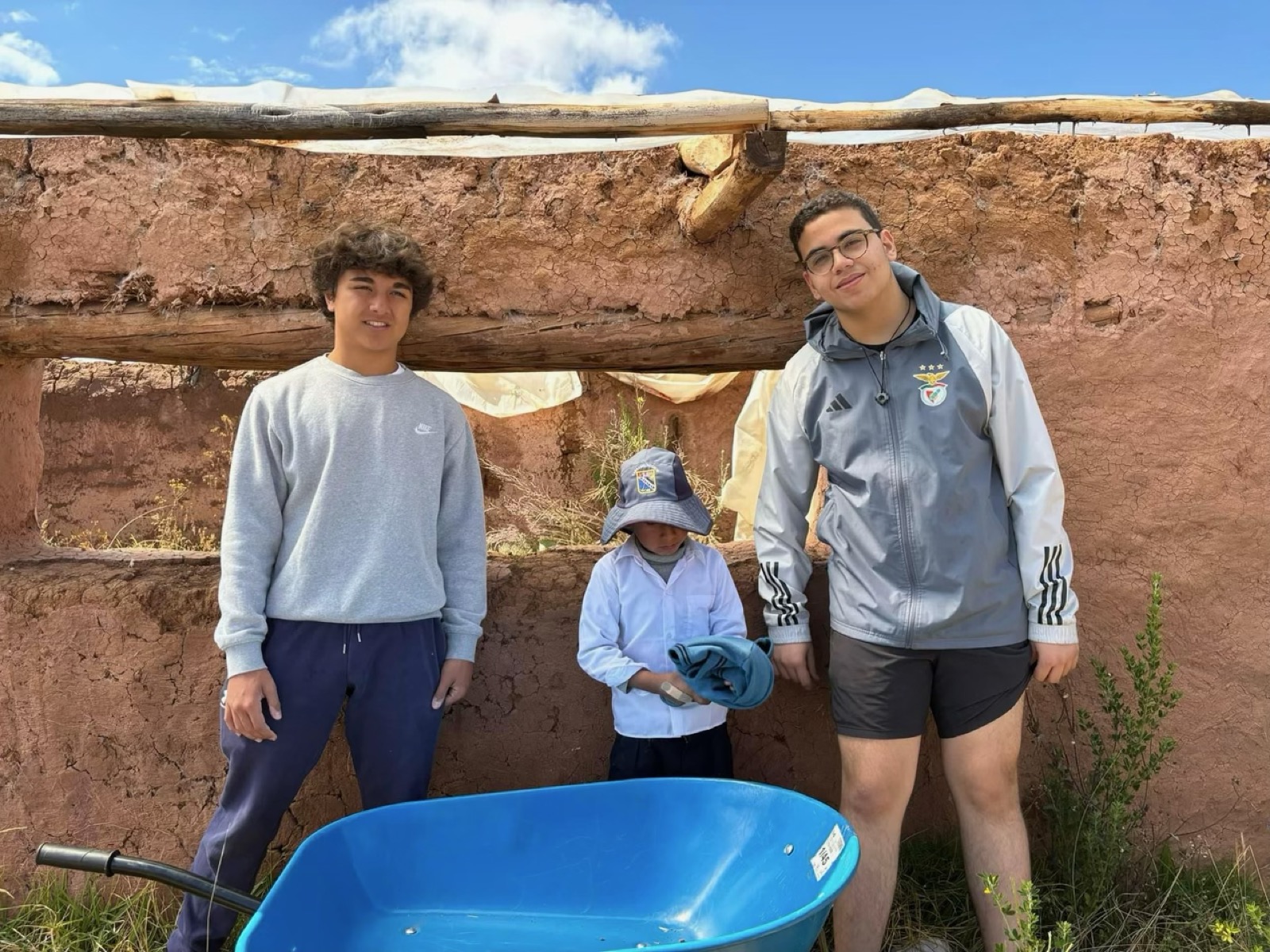 Two students next to a blue wheelbarrow in front of an adobe wall on a farm